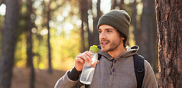 Ein Mann, bekleidet mit einem Hoodie und einer Mütze, steht in einem Wald. Die Sonne scheint und er setzt eine Wasserflasche zum Trinken an.