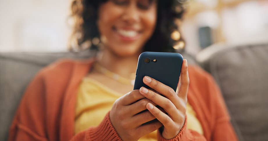 Eine junge Frau sitzt auf einem Sofa, hält ein Smartphone in ihrer Hand und schaut darauf. Das Smartphone ist auf dem Bild im Fokus.