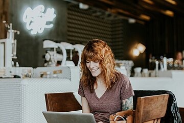 Eine junge, tätowierte Frau mit roten Haaren sitzt in einem Café und lächelt in ihrem Laptop.