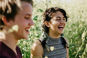 Ein junger Mann (vorne links im Bild, unscharf) und eine junge Frau (im Fokus) laufen gemeinsam durch ein Kornfeld und lachen herzhaft.