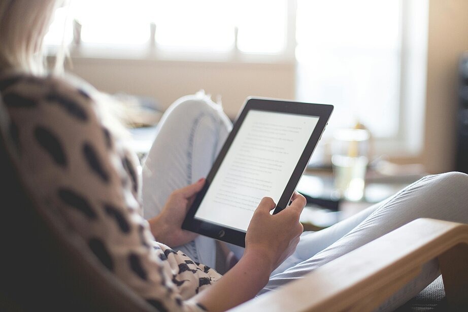 Zu sehen ist eine Frau, die mit einem Tablet auf einem Stuhl sitzt und liest. Sie wird schräg von hinten aufgenommen und sitzt im Schneidersitz auf dem Stuhl. Ihr Kopf ist nicht zu sehen, das Tablet und ihre Hände sind im Fokus.