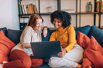 Zu sehen sind zwei junge Frauen, die gemütlich auf einem Sofa sitzen und gemeinsam lachend in einen Laptop schauen.